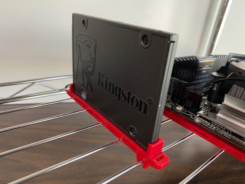 May include: A black Kingston solid state drive (SSD) with a red mounting bracket. The SSD is mounted to a metal shelf.