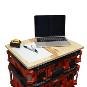 May include: A stack of two red toolboxes with black handles and latches. A laptop computer and a sheet of paper with two pens and a tape measure are on top of a wooden board placed on the toolboxes.