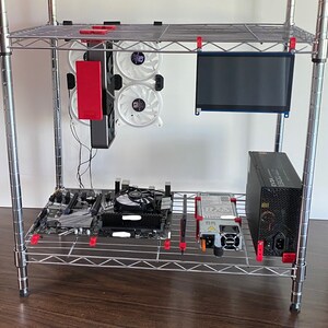 May include: A silver metal shelf unit with a computer motherboard, a power supply, a small monitor, and three white computer fans. The fans are attached to a black metal bracket.