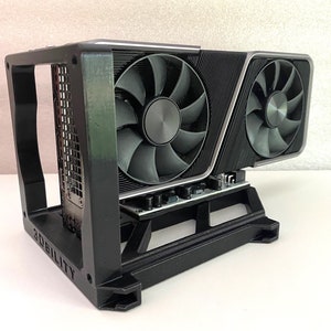 May include: A black graphics card with two fans is mounted in a black 3D printed stand. The stand has the word "2Dbility" printed on the side.