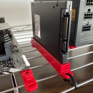 May include: A black SSD hard drive with a red 3D printed bracket and a black cable tie. The hard drive is mounted on a silver wire rack.