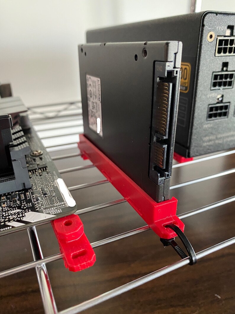 May include: A black SSD hard drive with a red 3D printed bracket and a black cable tie. The hard drive is mounted on a silver wire rack.
