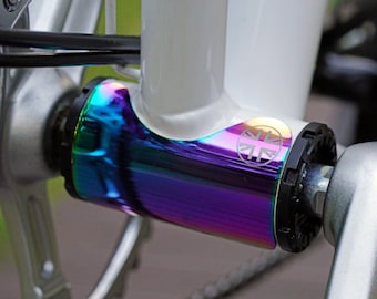 May include: A bicycle bottom bracket shell featuring an iridescent oil slick finish with a circular logo depicting a stylized Union Jack flag. The component is attached to a white bicycle frame and is positioned between a silver crank arm and chainring.