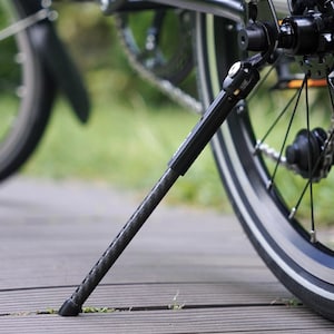 May include: A black bicycle kickstand with a carbon fibre texture on the lower leg, mounted to the rear frame near the wheel hub. The kickstand features a metallic hinge mechanism and a black rubber foot at the base to provide stability for the bike.