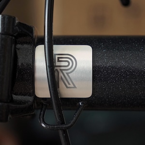 Peut inclure: Gros plan sur un cadre de vélo avec une plaque métallique argentée. La plaque présente une lettre "R" stylisée en contour gris foncé. Le cadre est noir avec une texture mouchetée, et un câble noir est visible.