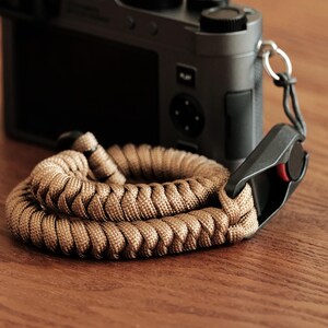 Może przedstawiać: Pasek do aparatu z linki paracord w kolorze opalenizny o plecionym wzorze, wyposażony w czarne plastikowe szybkozłącze z małym czerwonym okrągłym akcentem, przymocowany do szarego korpusu aparatu.