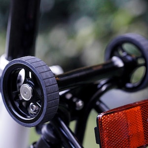 Puede incluir: Primer plano de un estabilizador de rueda de bicicleta negro con dos pequeñas ruedas negras. Las ruedas tienen un diseño de neumático ranurado. Un reflector rojo es visible. El cuadro de la bicicleta es blanco y negro.