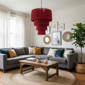 May include: A tiered, deep red fringe chandelier hangs above a gray sectional sofa. The sofa features teal and mustard yellow throw pillows. A rectangular wooden coffee table sits on a round jute rug, holding a book, a small plant, and a mug.