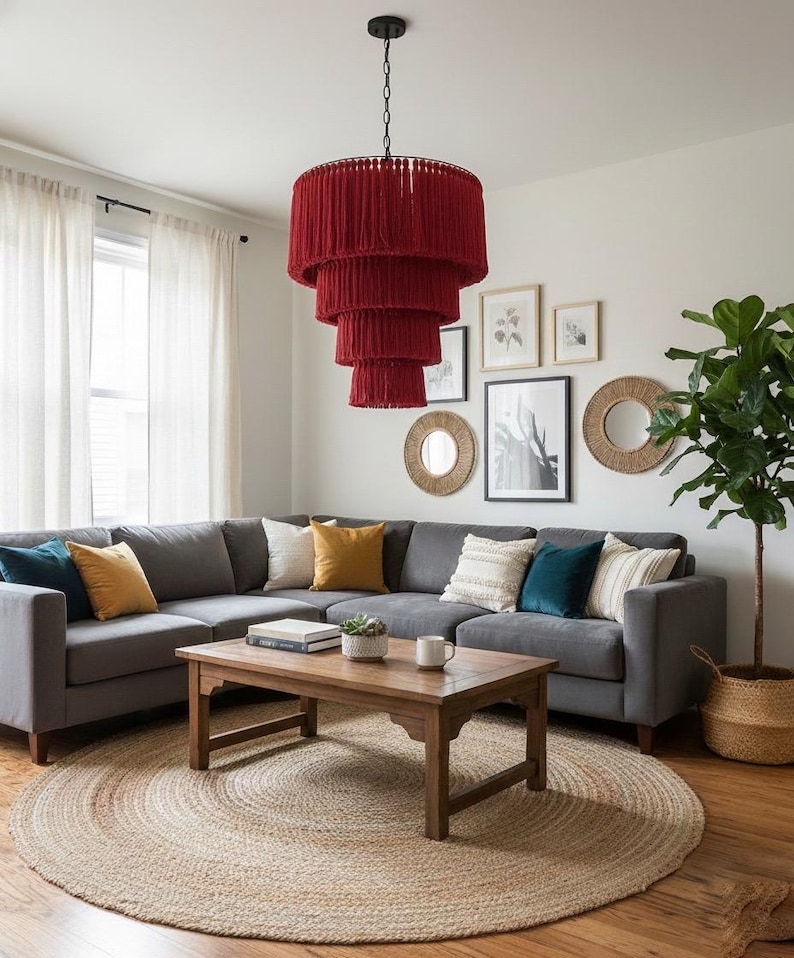 May include: A tiered, deep red fringe chandelier hangs above a gray sectional sofa. The sofa features teal and mustard yellow throw pillows. A rectangular wooden coffee table sits on a round jute rug, holding a book, a small plant, and a mug.