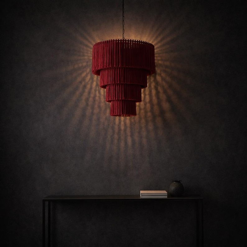 May include: A tiered pendant light fixture featuring four concentric circular layers of deep red fringe. The lamp is suspended by a thin chain and emits a warm, glowing light that casts a radial pattern of shadows against the surrounding wall space.