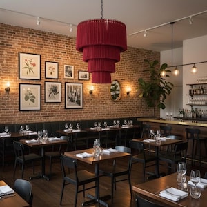 May include: A large, tiered, deep red fringe chandelier hangs above a restaurant dining area. The space features dark wood tables, black chairs, a brick wall adorned with framed botanical prints, and a bar area with glassware.