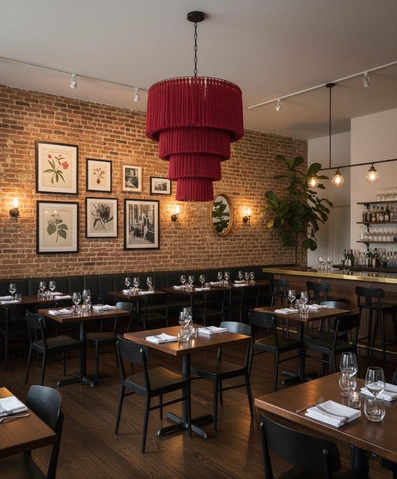 May include: A large, tiered, deep red fringe chandelier hangs above a restaurant dining area. The space features dark wood tables, black chairs, a brick wall adorned with framed botanical prints, and a bar area with glassware.
