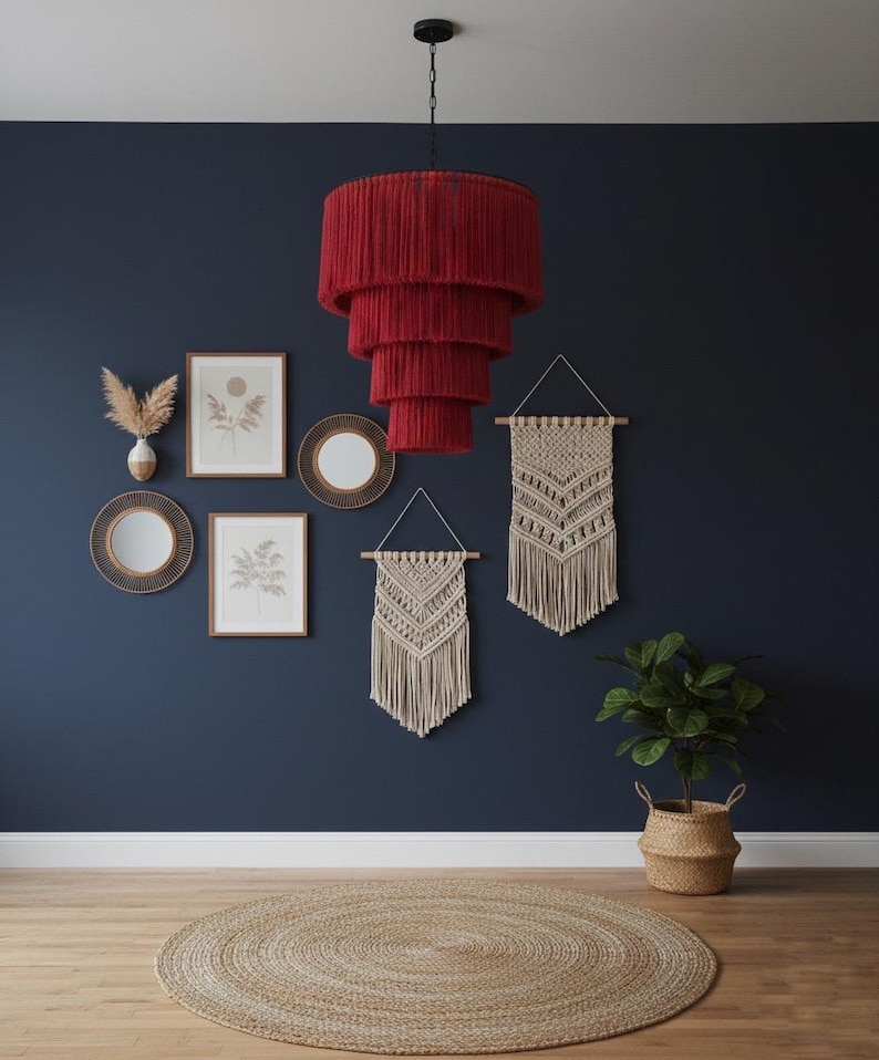 May include: A tiered, deep red fringe chandelier hangs from a black ceiling mount. The fixture features four concentric circular layers of red yarn, creating a cascading, cylindrical shape that provides a bold focal point in the room.