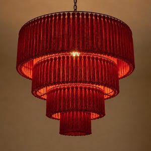 May include: A four-tiered chandelier featuring a circular frame adorned with dense, vibrant red fringe tassels. The structure is suspended by a thin metal chain, with a single light source glowing from within the center of the layered red fabric.