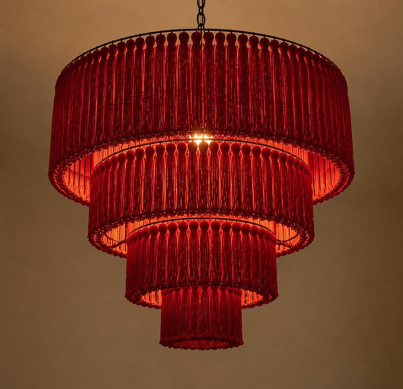 May include: A four-tiered chandelier featuring a circular frame adorned with dense, vibrant red fringe tassels. The structure is suspended by a thin metal chain, with a single light source glowing from within the center of the layered red fabric.