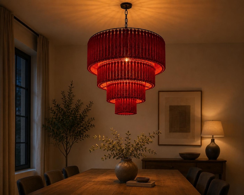 May include: A tiered chandelier featuring four concentric circular layers of deep red fringe. The fixture is suspended by a dark metal chain and emits a warm, glowing light that illuminates the surrounding space from within the layered fabric.