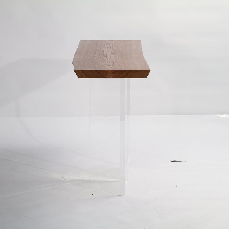 Puede incluir: Una mesa consola de madera con una tapa de borde vivo y patas de acr&iacute;lico transparente. La mesa est&aacute; hecha de madera natural con un acabado marr&oacute;n claro.