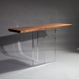 Puede incluir: Una mesa consola de madera con un acabado de madera natural y patas de acr&iacute;lico transparente. La mesa tiene una tapa rectangular y un dise&ntilde;o moderno.