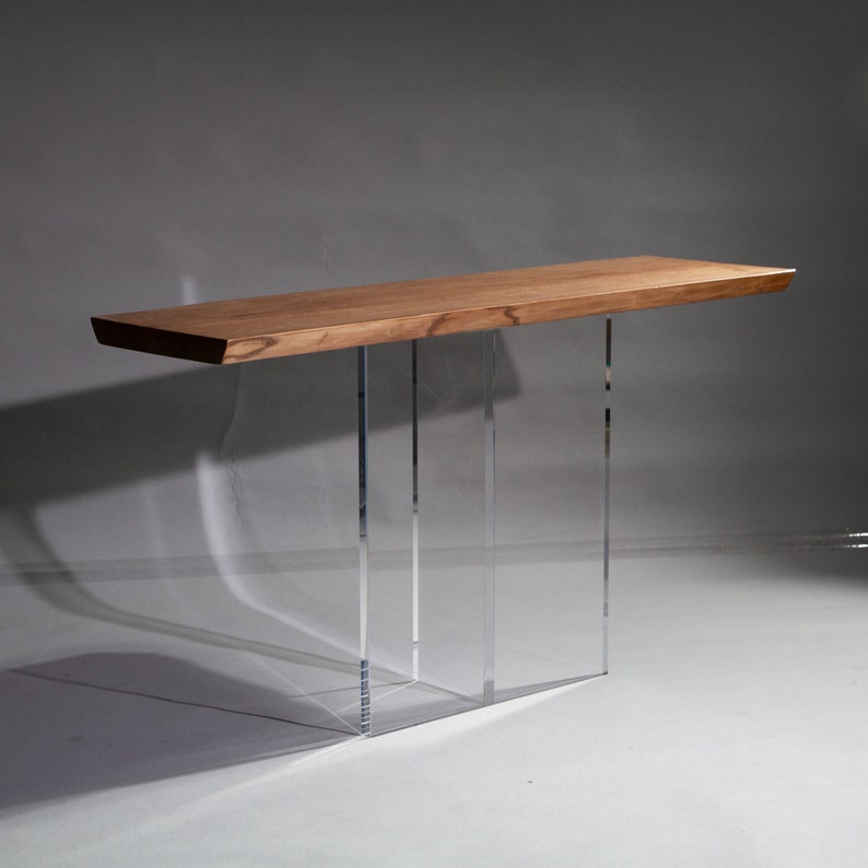 Puede incluir: Una mesa consola de madera con un acabado de madera natural y patas de acr&iacute;lico transparente. La mesa tiene una tapa rectangular y un dise&ntilde;o moderno.