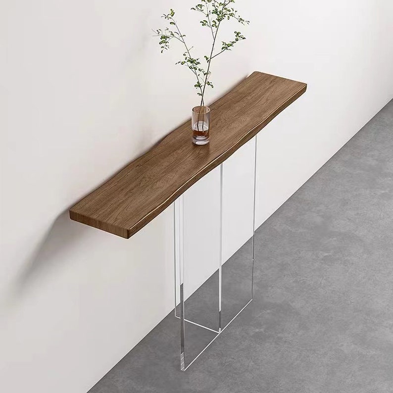 Puede incluir: Una mesa consola de madera con una tapa de borde vivo y patas de acr&iacute;lico transparente. La mesa est&aacute; montada en una pared blanca y tiene un peque&ntilde;o jarr&oacute;n con ramas verdes en la parte superior.