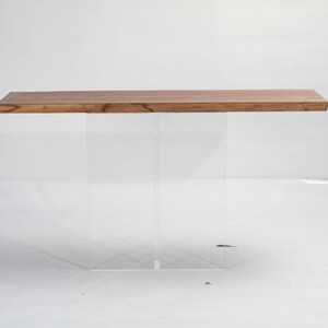 Puede incluir: Una mesa de madera rectangular larga con una base de acr&iacute;lico transparente. La mesa tiene un acabado de madera natural y la base est&aacute; hecha de dos patas de acr&iacute;lico transparente.