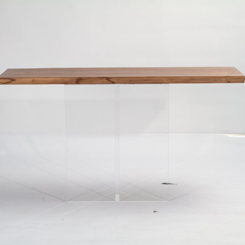 Puede incluir: Una mesa de madera rectangular larga con una base de acr&iacute;lico transparente. La mesa tiene un acabado de madera natural y la base est&aacute; hecha de dos patas de acr&iacute;lico transparente.