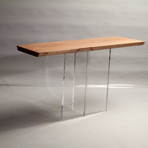Puede incluir: Una mesa consola larga de madera con un acabado de madera natural y patas de acr&iacute;lico transparente. La mesa tiene un dise&ntilde;o simple y moderno.