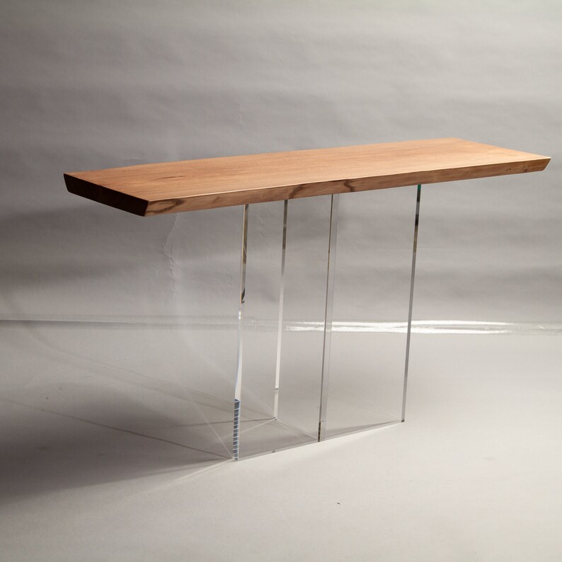 Puede incluir: Una mesa consola larga de madera con un acabado de madera natural y patas de acr&iacute;lico transparente. La mesa tiene un dise&ntilde;o simple y moderno.