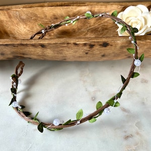 May include: A brown vine wreath with green leaves and white pearl beads. The wreath is lying on a light brown surface.