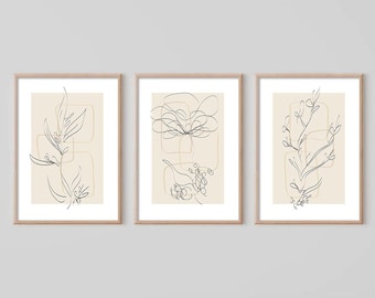 Puede incluir: Conjunto de tres impresiones minimalistas de arte lineal con diseños florales abstractos en negro y dorado sobre un fondo crema. Cada impresión está enmarcada en un marco de madera claro.