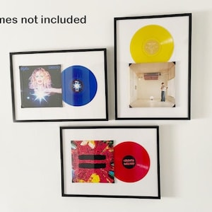 May include: Three framed vinyl records on a white wall. The top frame features a yellow record with a white label. The middle frame has a blue record with a woman on the album cover. The bottom frame has a red record with a colourful abstract design on the album cover. Frames not included.