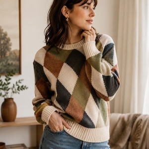 May include: A long-sleeve sweater featuring a multi-colored argyle pattern in shades of cream, olive green, dark gray, and rust brown. The knit garment has a crew neckline and ribbed cuffs and hem, paired with classic blue denim jeans.