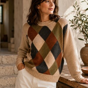 May include: A long-sleeve knit sweater featuring a beige base color with a prominent argyle pattern across the front. The diamond-shaped design incorporates earthy tones of dark charcoal, olive green, and rust brown, finished with a ribbed crew neckline.