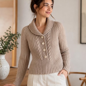 May include: A beige knitted cardigan with a shawl collar and button closure. The sweater features a cable knit design on the front and is paired with white pants. The woman is standing near a wooden table and a vase with greenery.