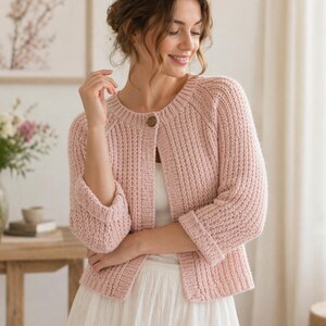 May include: A light pink, ribbed knit cardigan featuring a round neckline, three-quarter length sleeves with folded cuffs, and a single brown button closure at the top. The garment is worn over a simple white top, showcasing a textured, vertical knit pattern.