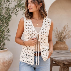 May include: A cream-coloured crochet vest with a tie closure. The open-weave design features a scalloped edge and a V-neck. The vest is worn over a neutral-coloured top and paired with blue jeans. A brown belt is visible.