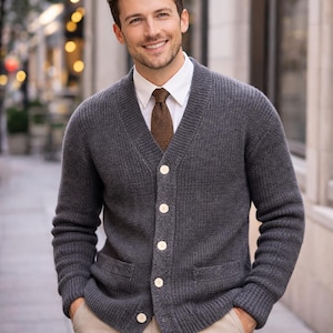 Può includere: Un cardigan grigio antracite con scollo a V, chiusura con bottoni e due tasche anteriori. Il maglione è indossato sopra una camicia bianca con colletto e una cravatta marrone. I bottoni sono color crema chiaro.