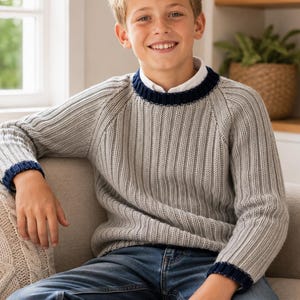 May include: A light grey long-sleeve jumper with a ribbed knit texture, featuring a dark navy blue trim along the crew neckline and sleeve cuffs, worn over a white collared shirt paired with blue denim jeans.