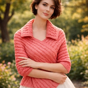 May include: A coral-colored knitted wrap sweater with a textured cable knit design. The sweater features a shawl collar and three-quarter sleeves. The wrap style creates a V-neckline. The background is a blurred outdoor scene with green and yellow foliage.