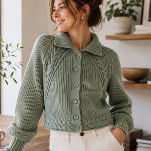 Könnte beinhalten: Eine salbeigrüne Strickjacke mit Kragen, langen Ärmeln mit gerippten Bündchen und einer durchgehenden Knopfleiste. Das Kleidungsstück zeigt vertikale Rippenmuster an Körper und Ärmeln, ergänzt durch filigrane Zopfstrickdetails an den Schultern.