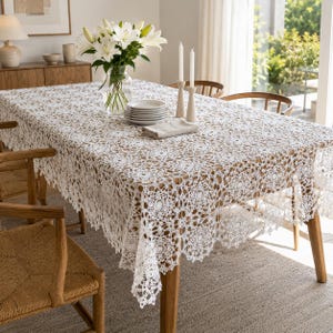 May include: A white rectangular tablecloth featuring an intricate crochet lace pattern with floral motifs. The tablecloth drapes over a wooden dining table, which is set with a stack of white ceramic plates, a folded napkin, and two candles.