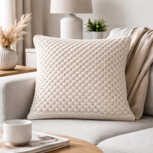 May include: A square throw cushion featuring a cream-coloured knit fabric with a raised, repeating honeycomb-like texture. The cushion has a solid, uniform appearance with a clean, finished edge, providing a soft and tactile surface for home decor.
