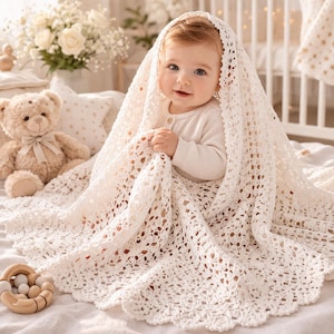 May include: A cream-colored, intricate crochet baby blanket with a scalloped edge draped over a baby wearing a long-sleeved white shirt. The blanket features a repeating openwork pattern and is made of soft yarn, providing a delicate texture.