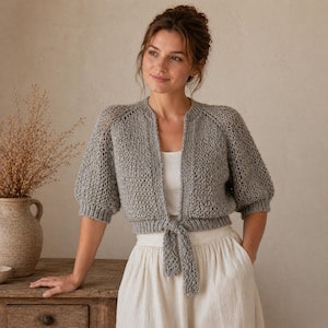 Peut inclure: Un gilet en tricot gris clair ouvert sur le devant, doté de manches courtes bouffantes et d'un lien à nouer à la taille. Le vêtement présente un motif en maille lâche et texturée avec des finitions côtelées aux poignets, à l'encolure et à l'ourlet.