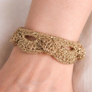 Könnte beinhalten: Ein goldfarbenes, gehäkeltes Armband mit offenem Design. Das Armband besteht aus metallischem Garn und hat ein strukturiertes Aussehen. Das Armband wird am Handgelenk getragen.
