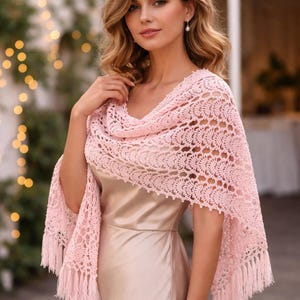 Puede incluir: Un chal de crochet rosa claro con un diseño floral y flecos. El chal está drapeado sobre un vestido. El diseño calado y el color suave crean un accesorio elegante, adecuado para ocasiones especiales o para añadir un toque de sofisticación a un atuendo.