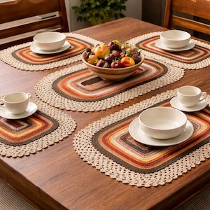 May include: A wooden table set with five crocheted placemats and matching coasters. The placemats feature a rectangular design with a gradient of brown, orange, and beige. White bowls and cups are placed on the mats, and a bowl of fruit sits in the center.
