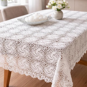 Puede incluir: Un mantel de encaje blanco y crochet cubre una mesa de madera. El intrincado patrón presenta diseños en espiral repetidos. Un plato de vidrio con esferas decorativas blancas y un jarrón con flores blancas se encuentran sobre la mesa.