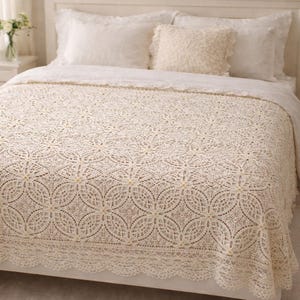 Op de afbeelding: Een crèmekleurig, gehaakt sprei met een gedetailleerd bloemmotief. De sprei is gedrapeerd over een bed met witte kussens en een bijpassend kussen. De sprei heeft een geschulpte kanten rand.