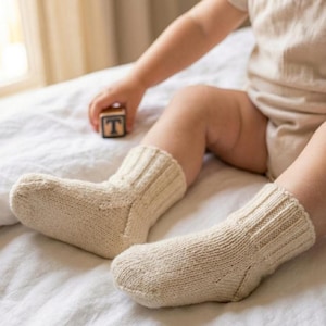 May include: A pair of cream-colored knitted baby socks. The socks have a ribbed cuff and a textured knit pattern. The socks are on a white surface. A wooden block with the letter "T" is in the background.